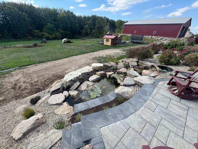 Scenic garden view with pond, stone patio, and a red barn in the background.
